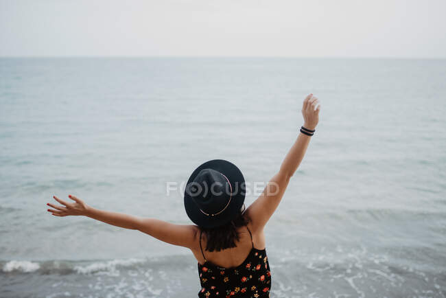 Visão traseira de alto ângulo da fêmea em chapéu preto e vestido de pé com braços levantados e desfrutando da vida na praia contra ondas oceânicas problemáticas em tempo nublado — Fotografia de Stock