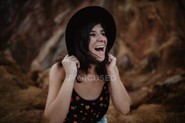 Happy confident stylish female in flowered shirt and denim shorts laughing looking away and holding black hat while standing against blurred brown rocky hill — Stock Photo