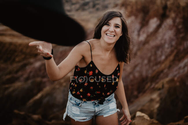 Счастливая уверенная стильная женщина в цветущей рубашке и джинсовых шортах смеется над камерой и бросает черную шляпу, стоя против размытого коричневого скалистого холма — стоковое фото