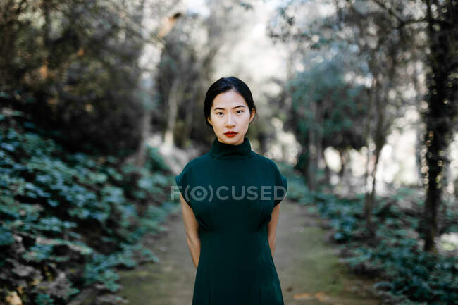 Молода азійка в модному одязі в зелених кущах і дивиться на камеру в старому саду. — стокове фото