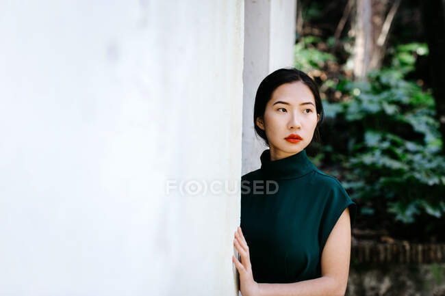 Junge Asiatin im trendigen Kleid im grünen Gebüsch und wegguckend an weiße Wand im betagten Garten gelehnt — Stockfoto