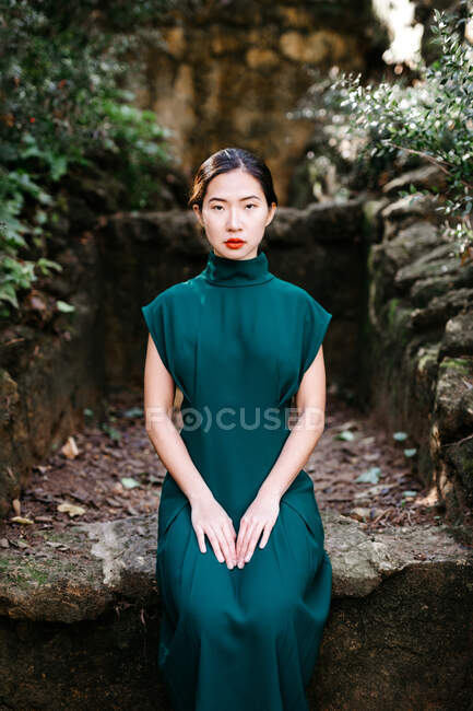 Jeune femme asiatique en robe à la mode assise sur une structure en pierre brute près des buissons verts et regardant la caméra dans le jardin âgé — Photo de stock