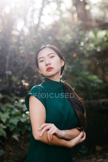 Молодая азиатка в модном платье в зеленых кустах и смотрит в камеру в старом саду — стоковое фото