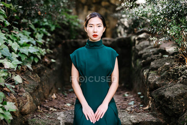 Jeune femme asiatique en robe à la mode assise sur une structure en pierre brute près des buissons verts et regardant la caméra dans le jardin âgé — Photo de stock