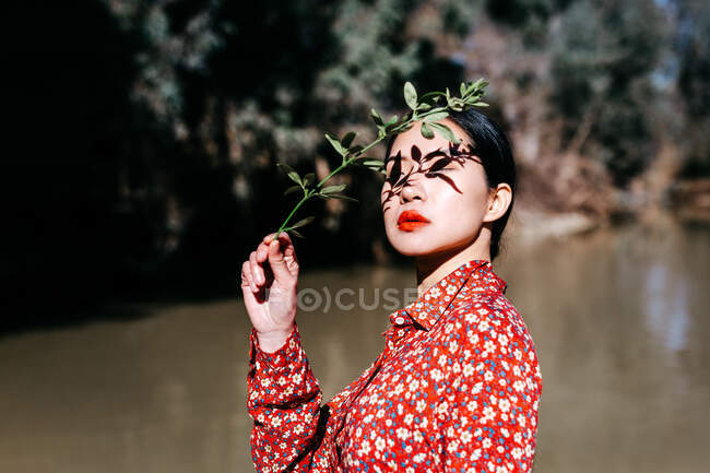Belle femme asiatique avec les yeux fermés et l'ombre de brindille de plante sur le visage debout contre le lac dans la campagne — Photo de stock