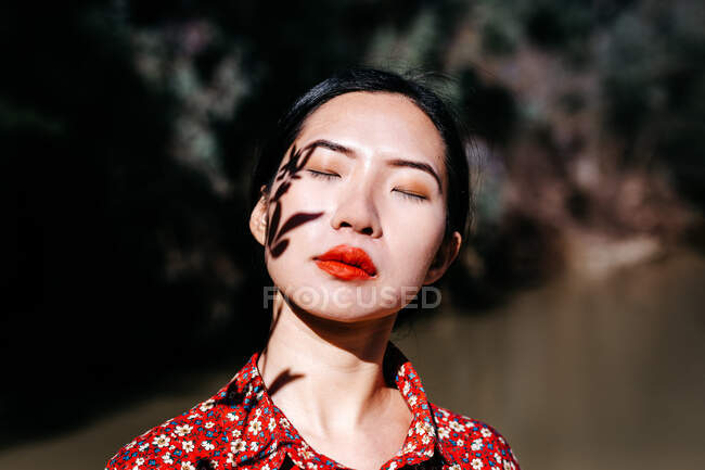 Красива азіатська жінка з закритими очима і тіні рослинної гілки на обличчі, стоячи проти озера в сільській місцевості — стокове фото