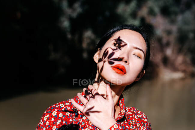 Belle femme asiatique avec les yeux fermés et l'ombre de brindille de plante sur le visage debout contre le lac dans la campagne — Photo de stock