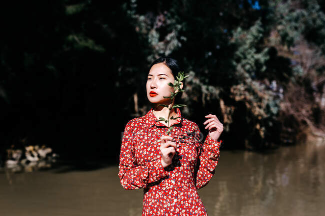 Beautiful asian woman with closed eyes and shadow of plant twig on face standing against lake in countryside — Stock Photo