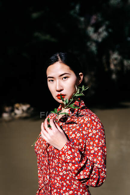 Bella donna asiatica guardando macchina fotografica in possesso di pianta ramoscello in piedi contro il lago in campagna — Foto stock