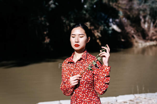 Stylish asian woman with closed eyes holding thin branch standing near peaceful lake on cloudless day in countryside — Stock Photo