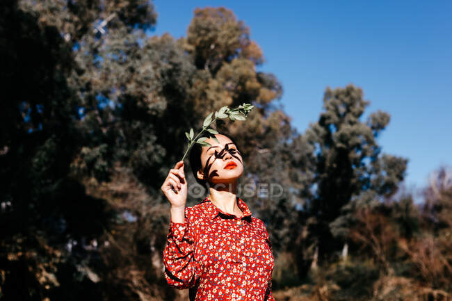 Elegante donna asiatica con gli occhi chiusi tenendo sottile ramo in braccia sollevate, mentre in piedi vicino al tranquillo lago il giorno senza nuvole in campagna — Foto stock