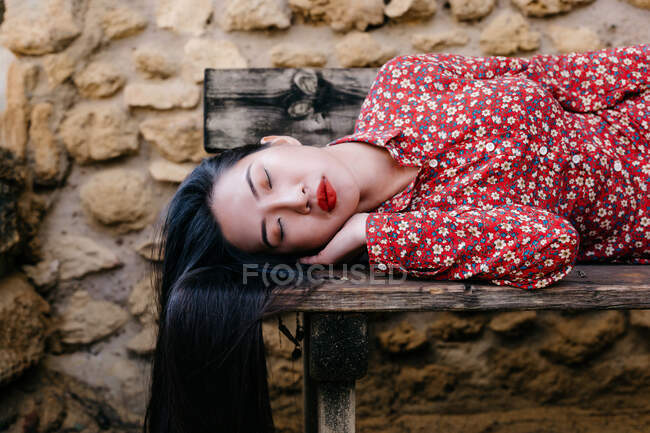 Mujer asiática en ropa floral de moda acostada en un banco de madera vieja con los ojos cerrados contra la pared de piedra - foto de stock