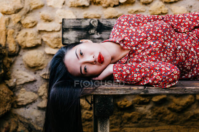 Азиатка в модной цветочной одежде лежит на старой деревянной скамейке и смотрит в камеру на каменную стену — стоковое фото