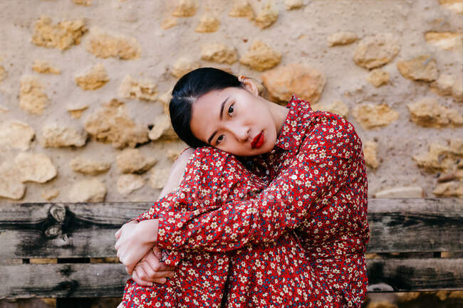 Seitenansicht der attraktiven asiatischen Frau im floralen Kleid, die Knie umarmt und in die Kamera schaut, während sie auf einer schäbigen Holzbank an einer Steinmauer sitzt — Stockfoto