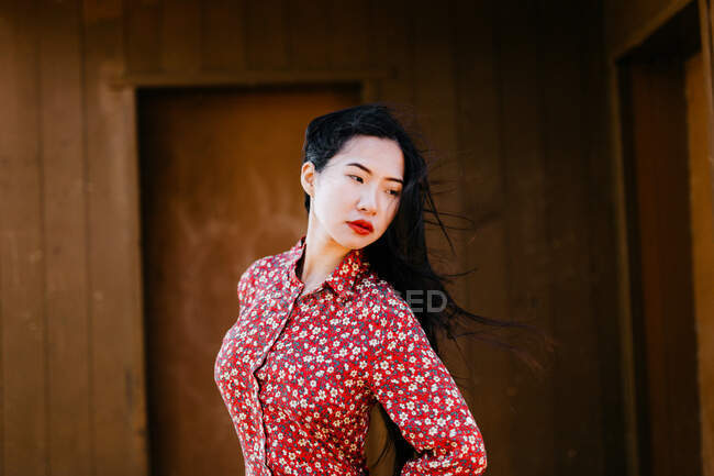 Attraktive asiatische Frau in stilvollem floralen Outfit schaut weg, während sie an windigen Tagen vor dem Holzhaus steht — Stockfoto