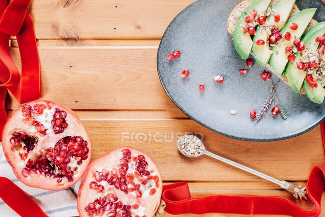 Blick von oben auf halbierten Granatapfel und Löffel mit Kernen in der Nähe des Tellers mit köstlichem Avocado-Toast auf Holztisch — Stockfoto