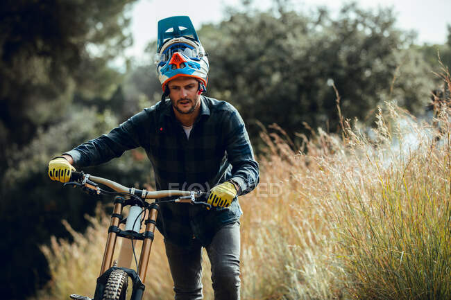 Bell'uomo in casco che cammina guardando giù mentre porta in sella la mountain bike dopo la pratica nel prato — Foto stock