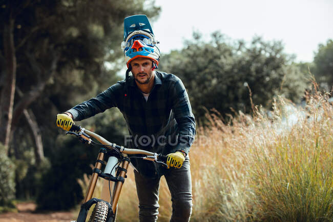 Beau homme en casque marchant en regardant la caméra tout en portant le vélo de montagne après l'entraînement dans la prairie — Photo de stock