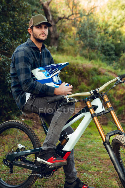 Desportista de bicicleta de montanha sem proteção segurando um capacete sentado em bicicleta no meio de uma floresta olhando para longe — Fotografia de Stock