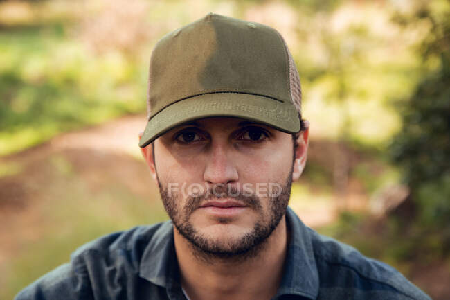 Retrato de hombre morena guapo en camisa a cuadros y gorra de béisbol de pie en el fondo de la naturaleza mirando a la cámara - foto de stock
