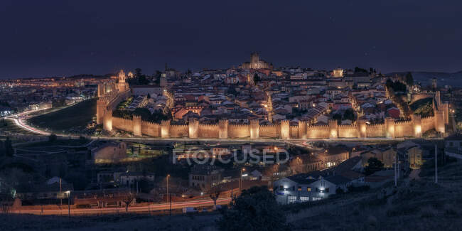 From above of medieval castle surrounded illuminated stone defensive walls with golden lights and contemporary night city under cloudy gray sky in Avila — Stock Photo
