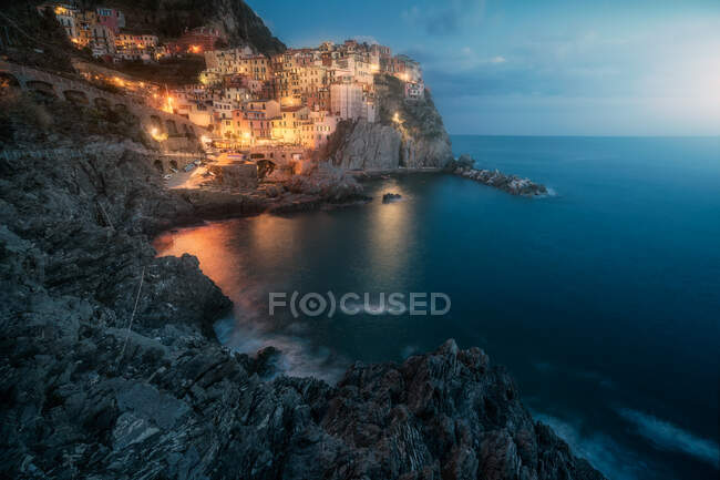 Paesaggio incredibile con piccola città con luci colorate sulla costa rocciosa lavaggio da acqua tranquilla oceano di notte — Foto stock