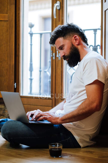 Вид збоку бородатий чоловік сидить на підлозі біля чашки гарячого напою і друкує на ноутбуці під час роботи над віддаленим проектом вдома — стокове фото