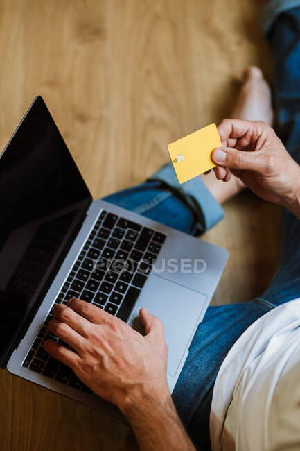 From above anonymous man with credit card sitting on floor and using laptop to make online purchases at home — Stock Photo