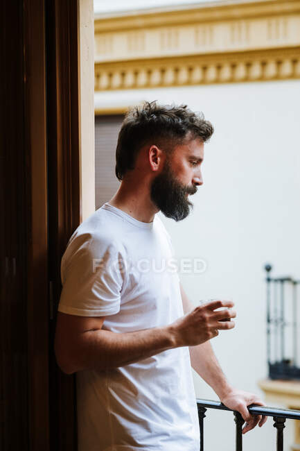 Seitenansicht eines bärtigen Mannes im weißen T-Shirt, der morgens auf dem Balkon steht und Heißgetränk genießt — Stockfoto
