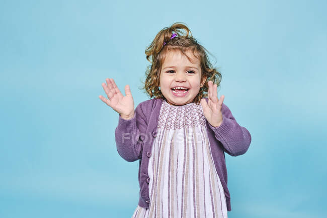 Alegre niño pequeño en vestido púrpura y cárdigan de punto sonriendo a la cámara y aplaudiendo mientras está parado solo contra el fondo azul claro en el estudio moderno - foto de stock