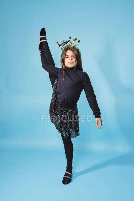 Trendy pleased child in New Year head wear and black clothes looking at camera and doing standing side split exercise against light blue background in studio — Stock Photo