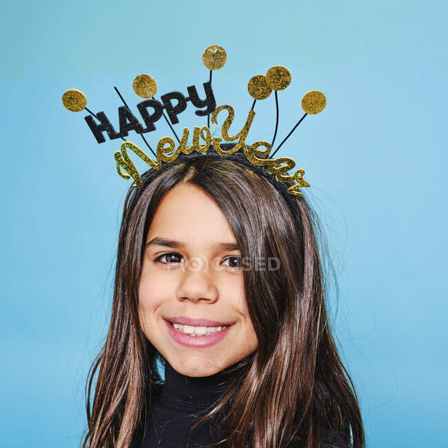 Happy preteen in black and gold headgear with wishes happy New Year smiling at camera against light blue background in modern studio — Stock Photo