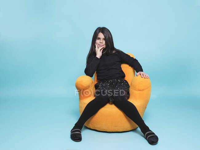 Hübsches Mädchen in schwarzer Freizeitkleidung, die Hand am Kinn liegend, in die Kamera blickend, während es sich in leuchtend gelben Sesseln vor hellblauem Hintergrund in einem modernen Studio entspannt — Stockfoto