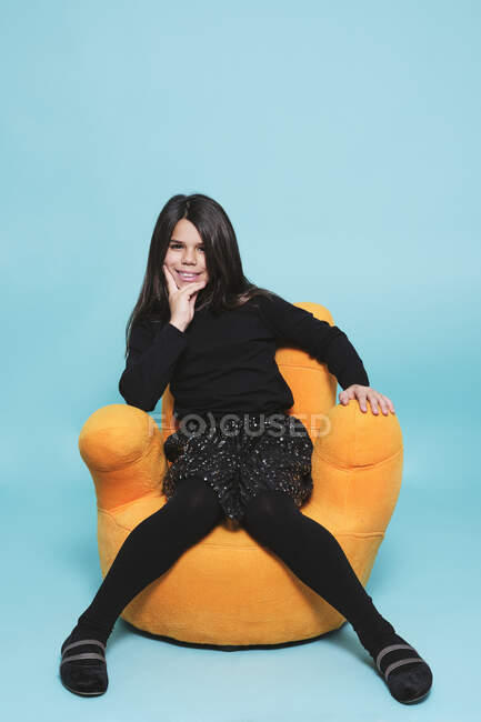 Hübsches Mädchen in schwarzer Freizeitkleidung, die Hand auf dem Kinn liegend lächelnd in die Kamera blickend, während es sich in leuchtend gelbem Sessel vor hellblauem Hintergrund in einem modernen Studio entspannt — Stockfoto