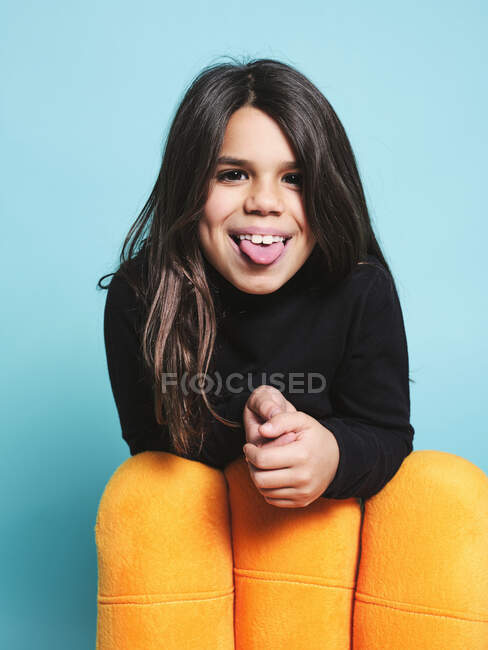 Menina pré-adolescente feliz em roupas casuais pretas sorrindo furando a língua para fora para a câmera enquanto se inclina atrás de poltrona amarela contra fundo azul claro no estúdio contemporâneo — Fotografia de Stock