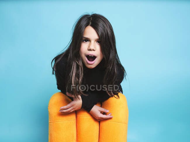 Happy preteen girl in black casual clothes smiling with mouth open looking at camera while leaning behind yellow armchair against light blue background in contemporary studio — Stock Photo