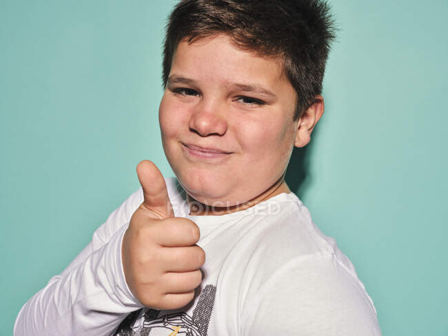 Joyful confident chubby plus size preteen boy in casual clothes looking at camera and laughing while gesturing thumbs up against turquoise background — Stock Photo