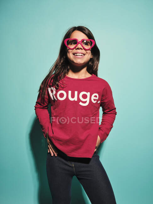Happy preteen girl in pink heart shape glasses and casual clothes smiling away while standing with hands on hips against turquoise background in modern studio — Stock Photo