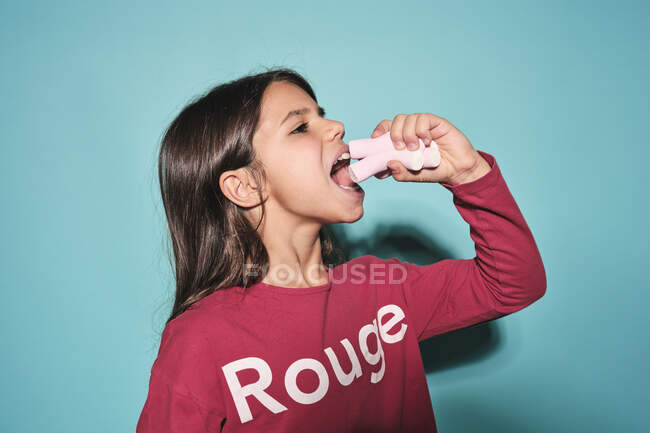 Vista lateral de la niña feliz en ropa casual roja comiendo malvavisco de caramelo suave rosa mirando hacia otro lado mientras está de pie sobre el fondo azul - foto de stock
