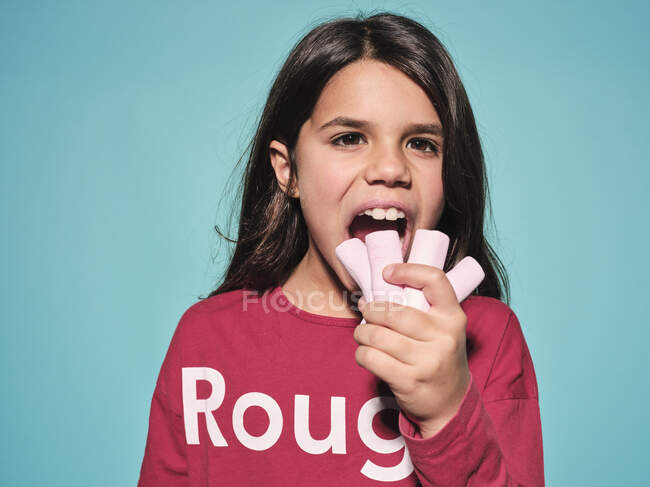 Happy little girl in red casual wear eating pink soft candy marshmallow looking away while standing against blue background — Stock Photo