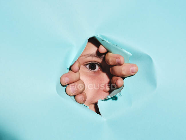 Niño curioso con ojos marrones mirando a la cámara a través de un agujero roto en papel azul brillante en el estudio - foto de stock