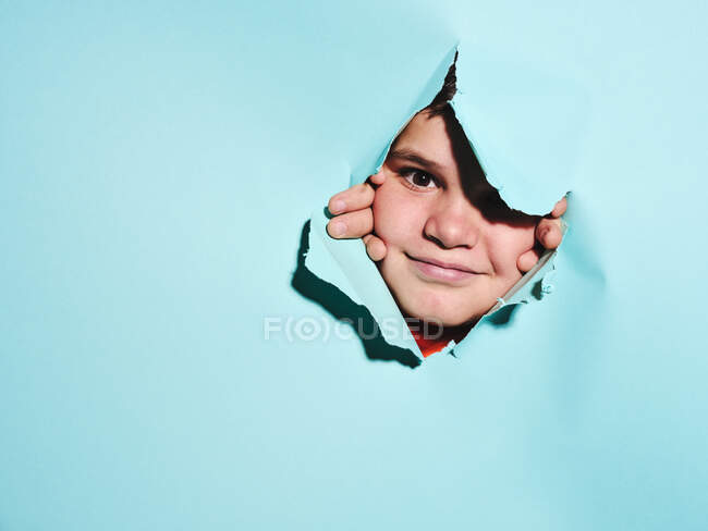 Fröhlich aufgeregter kleiner Junge mit Fratze blickt durch zerrissenes buntes blaues Papierblatt im Atelier in die Kamera — Stockfoto
