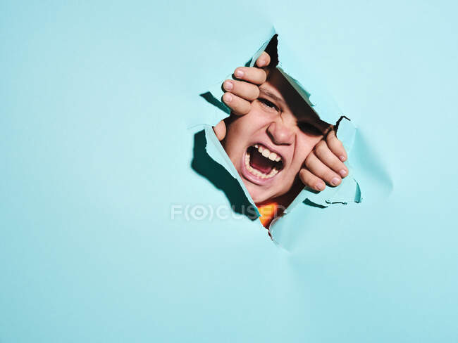 Кричащий злой мальчик с гримасой, смотрящий сквозь разорванную голубую бумажную простынь в студии — стоковое фото