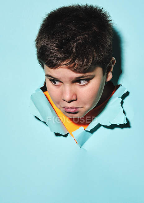 Cheerful excited little boy making funny faces looking away through ripped colorful blue paper sheet in studio — Stock Photo