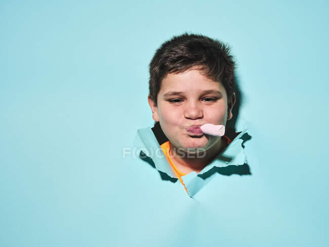 Menino gordinho feliz com doces rosa na boca sorrindo e olhando para a câmera através de papel rasgado azul — Fotografia de Stock