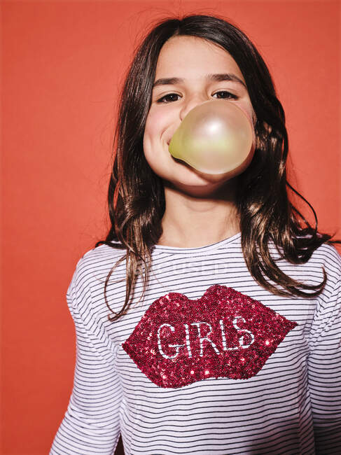 Cheerful preteen girl in trendy outfit blowing yellow bubble gum and looking at camera while standing against red background — Stock Photo