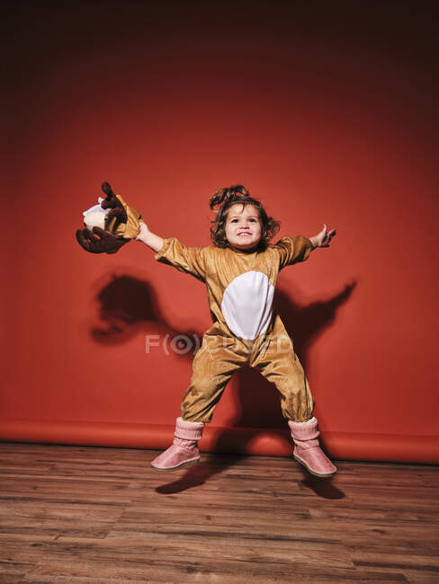 Energetic happy little girl in cute deer costume spreading arms while jumping looking up against red wall in studio — Stock Photo