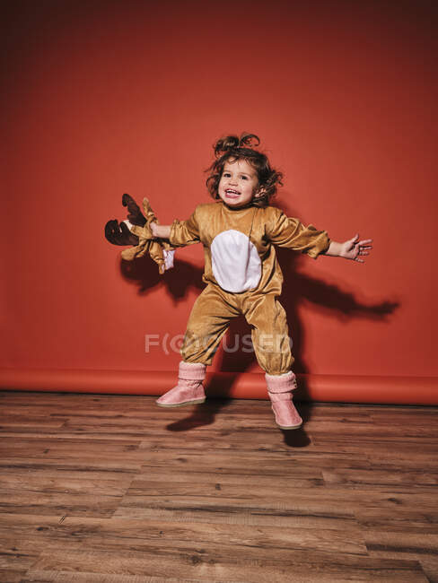 Енергійний щасливий маленька дівчинка в милий костюм оленя, розтягуючи руки, стрибаючи, дивлячись на червону стіну в студії — стокове фото