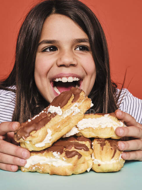 Close-up de menina alegre em desgaste casual desfrutando de doces eclairs com chocolate enquanto sentado à mesa contra o fundo vermelho — Fotografia de Stock