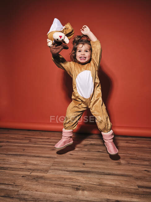Energique petite fille heureuse en costume de cerf mignon écartant les bras tout en sautant la recherche contre le mur rouge en studio — Photo de stock